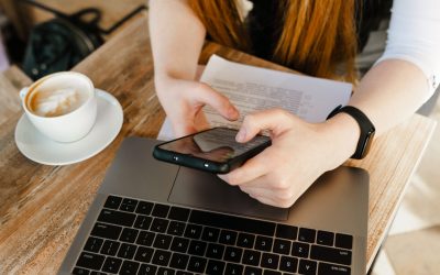Closeup photo of freelancer sitting at desk with laptop and business papers, using internet on smartphone wearing formal clothes. Girl iworks in a cafe with business papers and a laptop, holds phone
